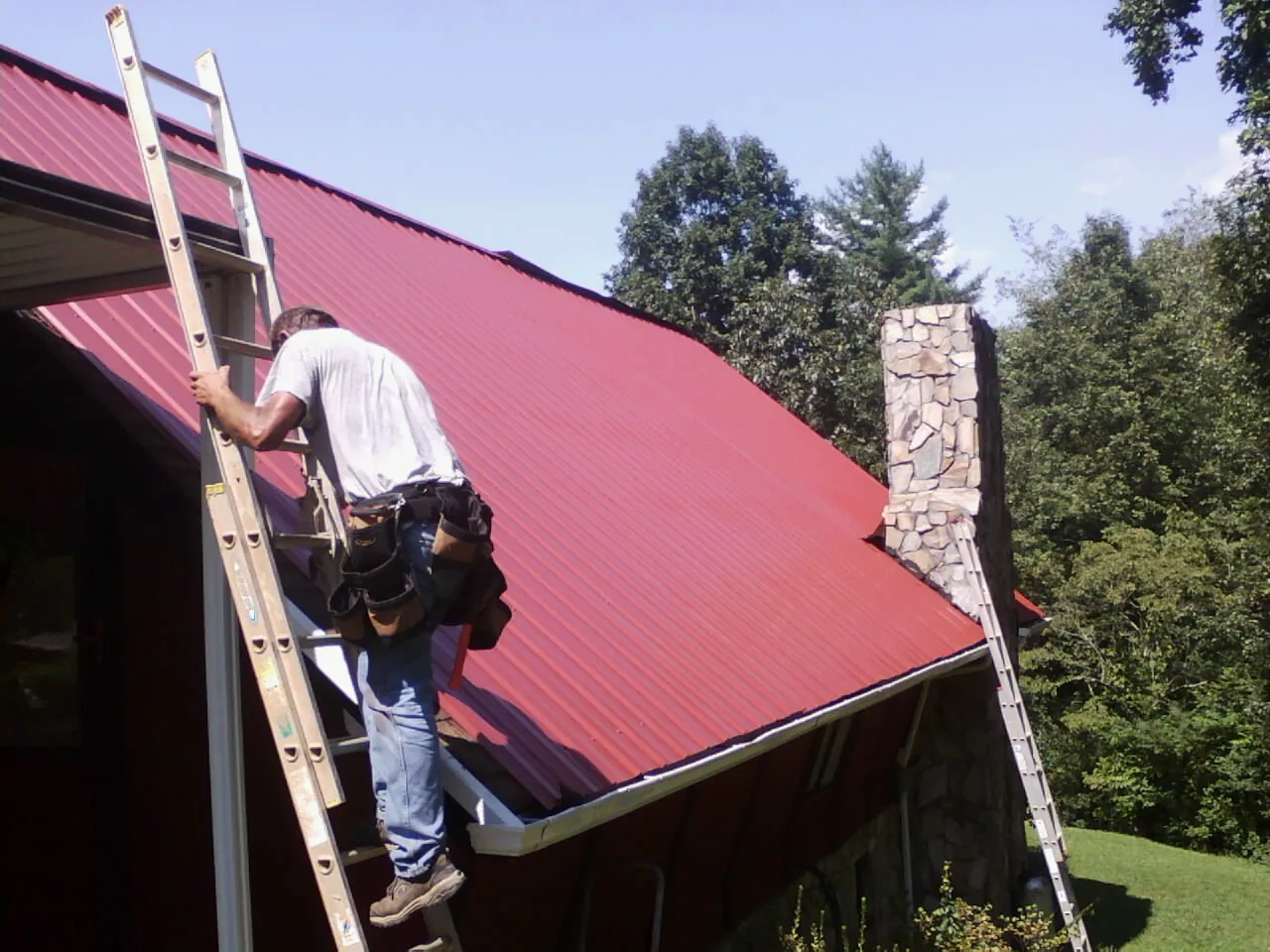 Roofer on a ladder beside a red metal roof and stone chimney — Soffit & Fascia Repair by Roofing Brothers in Timmonsville